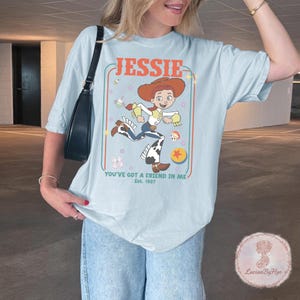 May include: Light blue t-shirt featuring a cartoon image of Jessie from Toy Story. The design includes the name "Jessie" in orange, the phrase "You've Got a Friend in Me," and the year "Est. 1997." The shirt has a relaxed fit.