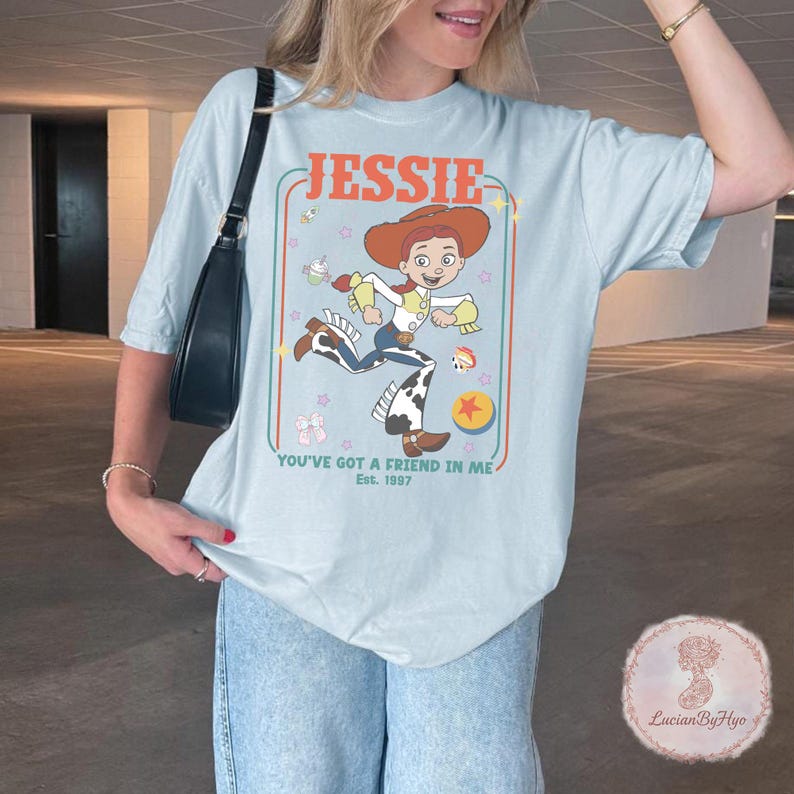 May include: Light blue t-shirt featuring a cartoon image of Jessie from Toy Story. The design includes the name "Jessie" in orange, the phrase "You've Got a Friend in Me," and the year "Est. 1997." The shirt has a relaxed fit.