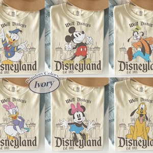 Puede incluir: Camisetas color marfil con varios personajes de Disney: Pato Donald, Mickey Mouse, Goofy, Daisy Duck, Minnie Mouse y Pluto. Cada camiseta muestra "Walt Disney's Disneyland Est. 1955" en un estilo vintage.