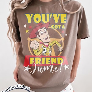 Op de afbeelding: Espresso-kleurig T-shirt met een Woody uit Toy Story-afbeelding. Het ontwerp bevat de tekst "YOU'VE GOT A FRIEND Jume!" in geel en wit, met sterren en een rode banner. Het shirt is van Comfort Colors.