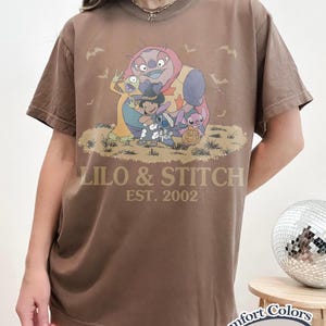 K&ouml;nnte beinhalten: Espressofarbenes T-Shirt mit einer Grafik von Lilo & Stitch-Charakteren in Halloween-Kost&uuml;men. Der Text "LILO & STITCH EST. 2002" ist auf dem Shirt aufgedruckt. Das Shirt ist von der Marke Comfort Colors.