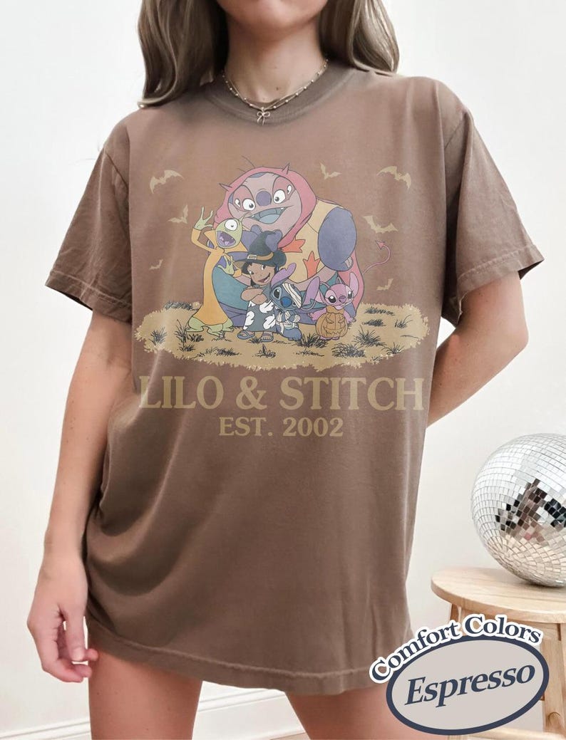 K&ouml;nnte beinhalten: Espressofarbenes T-Shirt mit einer Grafik von Lilo & Stitch-Charakteren in Halloween-Kost&uuml;men. Der Text "LILO & STITCH EST. 2002" ist auf dem Shirt aufgedruckt. Das Shirt ist von der Marke Comfort Colors.