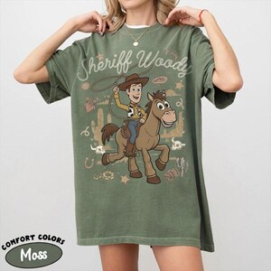 May include: A moss green short-sleeve t-shirt featuring a graphic of Sheriff Woody riding a brown horse. The design includes the text Sheriff Woody, a lasso, cacti, stars, and desert elements in a light tan color. The shirt has a crew neckline.