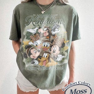 May include: Olive green t-shirt featuring a graphic of Disney characters, including Mickey and Minnie Mouse, Donald and Daisy Duck, and Goofy. The shirt has the words "Animal Kingdom" and "Walt Disney World" printed on it. The shirt is labeled "Comfort Colors Moss".