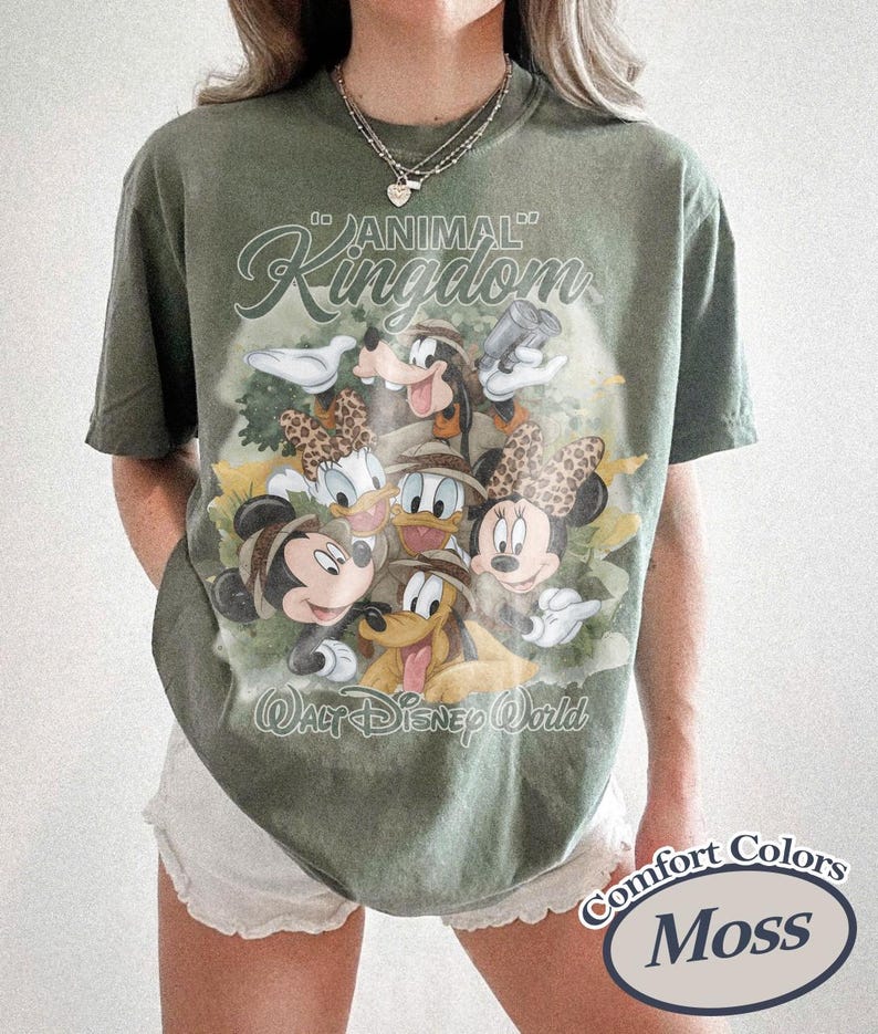 May include: Olive green t-shirt featuring a graphic of Disney characters, including Mickey and Minnie Mouse, Donald and Daisy Duck, and Goofy. The shirt has the words "Animal Kingdom" and "Walt Disney World" printed on it. The shirt is labeled "Comfort Colors Moss".