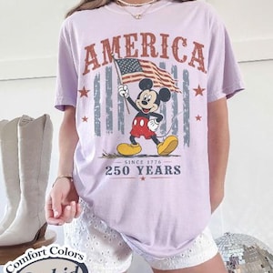Pu&ograve; includere: Una maglietta lilla chiaro con la scritta "AMERICA" in un font invecchiato. La maglietta presenta una grafica di Mickey Mouse che tiene una bandiera americana, con il testo "SINCE 1776" e "250 YEARS".