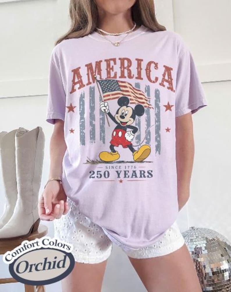 Pu&ograve; includere: Una maglietta lilla chiaro con la scritta "AMERICA" in un font invecchiato. La maglietta presenta una grafica di Mickey Mouse che tiene una bandiera americana, con il testo "SINCE 1776" e "250 YEARS".