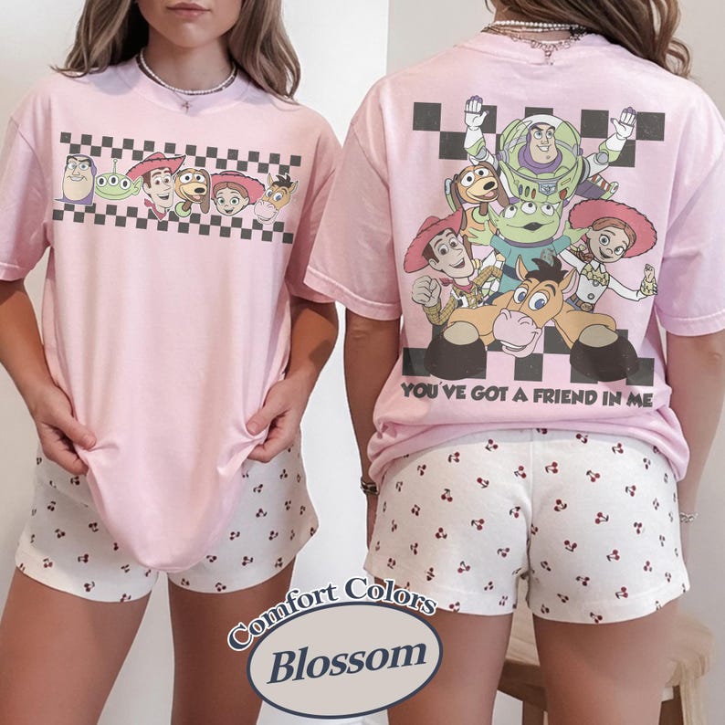 May include: Pink t-shirt and shorts set featuring Toy Story characters. The front of the shirt has a checkerboard design with cartoon characters. The back of the shirt has a larger graphic with the text "YOU'VE GOT A FRIEND IN ME". The shorts have a white background with a small cherry pattern.