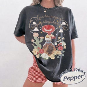 Peut inclure: Un t-shirt gris anthracite &agrave; manches courtes avec un motif repr&eacute;sentant une cow-girl au chapeau rouge, entour&eacute;e de cactus et de fleurs. Le texte indique Love has ways of bringing things back to life, avec des notes de musique et des nuages.