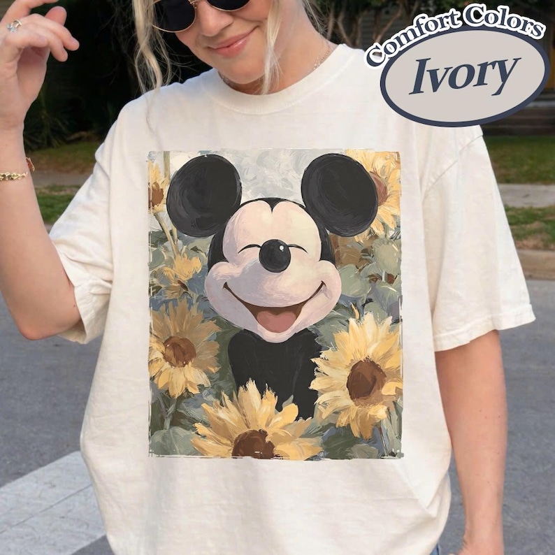 Puede incluir: Camiseta de color marfil con un dise&ntilde;o de Mickey Mouse sonriente rodeado de girasoles. El gr&aacute;fico es una ilustraci&oacute;n colorida con una est&eacute;tica vintage. La camiseta es de la marca Comfort Colors.