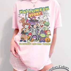 May include: A light pink t-shirt with a graphic featuring cartoon characters in Halloween costumes. The text "HALLOWEEN STORY" is above the characters, and "TO INFINITY AND BOO YOUND!" is below. The shirt is labeled "Comfort Colors Blossom".