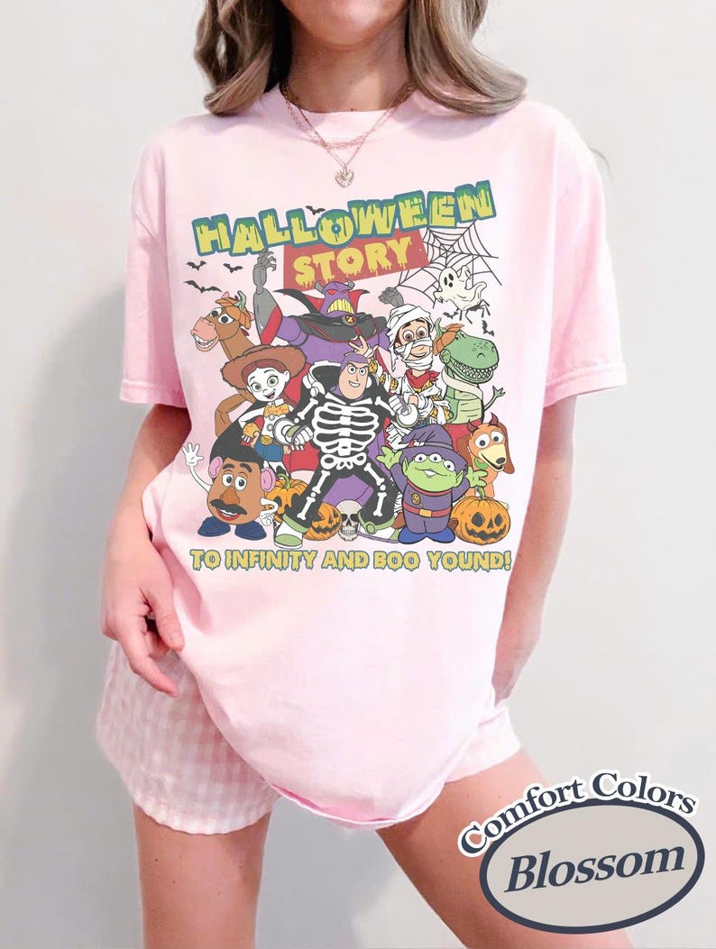May include: A light pink t-shirt with a graphic featuring cartoon characters in Halloween costumes. The text "HALLOWEEN STORY" is above the characters, and "TO INFINITY AND BOO YOUND!" is below. The shirt is labeled "Comfort Colors Blossom".