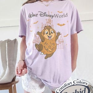 以下が含まれることがあります： 「ウォルト・ディズニー・ワールド」の文字と、カボチャのコスチュームを着たチップの漫画が描かれた薄紫色のTシャツ。デザインにはコウモリと城が含まれています。このシャツは「Comfort Colors Orchid」と表示されています。