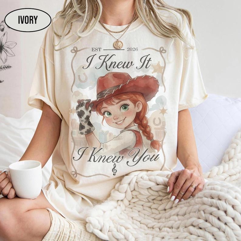 May include: An ivory short-sleeve t-shirt featuring a graphic of a cartoon cowgirl with red hair and a brown hat. The text reads "EST 2026", "I Knew It", and "I Knew You" in a decorative frame. The shirt has a crew neckline and a relaxed fit.