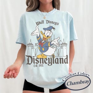 Puede incluir: Camiseta azul claro con Donald Duck con un traje de marinero azul marino y el texto "Walt Disney's Disneyland Est. 1955". La camiseta es Comfort Colors Chambray.