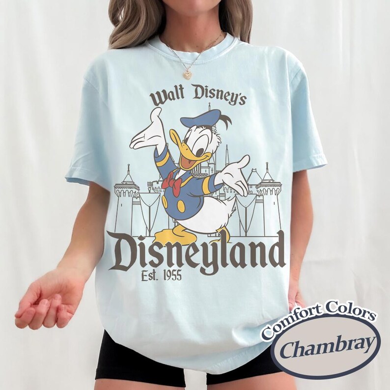 Puede incluir: Camiseta azul claro con Donald Duck con un traje de marinero azul marino y el texto "Walt Disney's Disneyland Est. 1955". La camiseta es Comfort Colors Chambray.