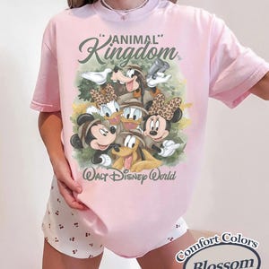 May include: A light pink t-shirt featuring a graphic of Disney characters, including Mickey and Minnie Mouse, Donald and Daisy Duck, and Goofy. The shirt has the words "Animal Kingdom" and "Walt Disney World" printed on it.