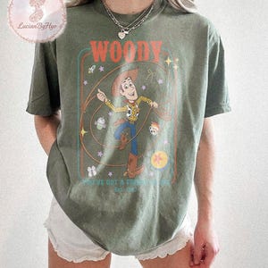 May include: Olive green t-shirt featuring a graphic of Woody from Toy Story. The design includes the word "WOODY" in orange, and the phrase "YOU'VE GOT A FRIEND IN ME" with the year "Est. 1995". The graphic is framed with a teal and orange border.