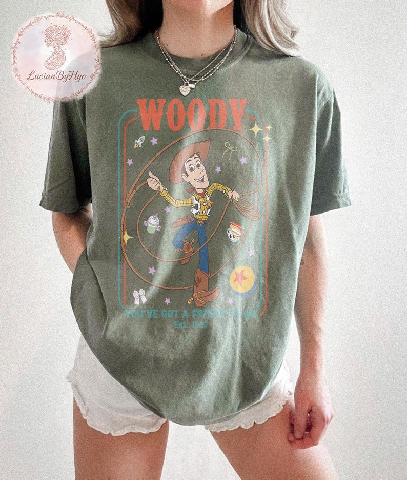 May include: Olive green t-shirt featuring a graphic of Woody from Toy Story. The design includes the word "WOODY" in orange, and the phrase "YOU'VE GOT A FRIEND IN ME" with the year "Est. 1995". The graphic is framed with a teal and orange border.