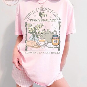 May include: A light pink t-shirt with a graphic featuring a teapot, teacup, honey jar, and flowers. The text reads "World Famous Gumbo, Tiana's Palace, Flower Tea Cake Honey."