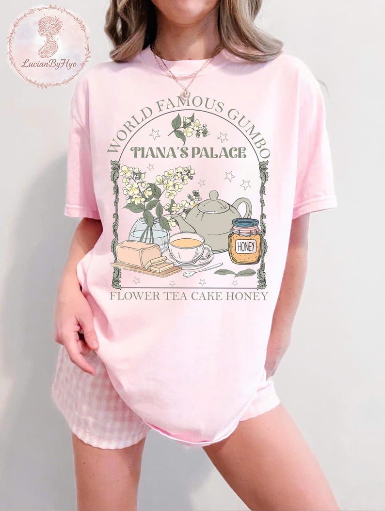 May include: A light pink t-shirt with a graphic featuring a teapot, teacup, honey jar, and flowers. The text reads "World Famous Gumbo, Tiana's Palace, Flower Tea Cake Honey."