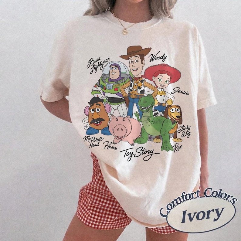 May include: Ivory-coloured t-shirt featuring a graphic of Toy Story characters including Woody, Buzz Lightyear, and Jessie. The shirt has the words "Toy Story" printed below the characters. The shirt is labelled "Comfort Colors" and "Ivory".