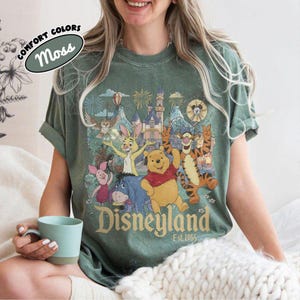 May include: A moss green short-sleeve t-shirt featuring a graphic print of Winnie the Pooh, Tigger, Piglet, Eeyore, and Rabbit in front of a castle with fireworks. The text reads Disneyland Est. 1955. A label in the corner says Comfort Colors Moss.