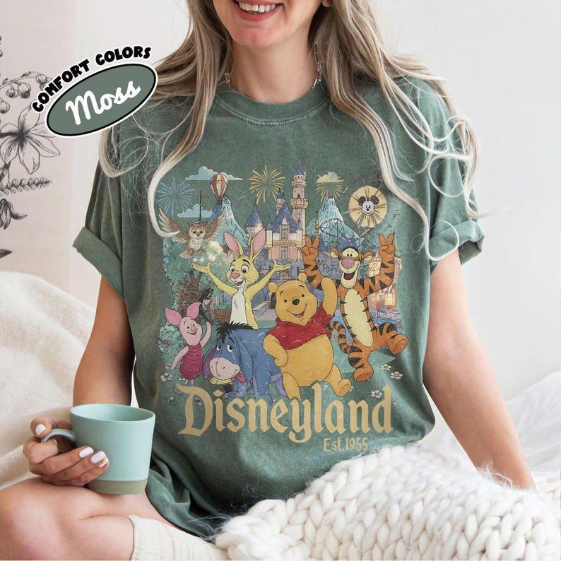 May include: A moss green short-sleeve t-shirt featuring a graphic print of Winnie the Pooh, Tigger, Piglet, Eeyore, and Rabbit in front of a castle with fireworks. The text reads Disneyland Est. 1955. A label in the corner says Comfort Colors Moss.