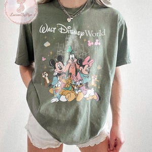 May include: Olive green t-shirt featuring a graphic of Disney characters, including Mickey Mouse, Minnie Mouse, and Donald Duck, with the text "Walt Disney World". The design includes a castle and other decorative elements.