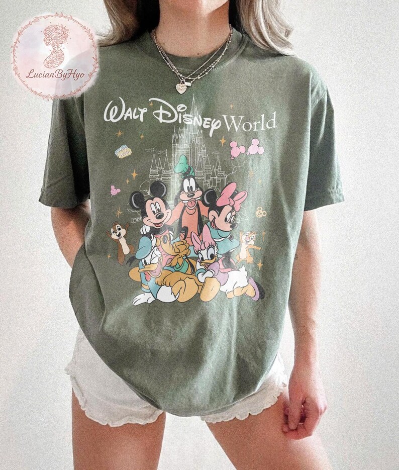 May include: Olive green t-shirt featuring a graphic of Disney characters, including Mickey Mouse, Minnie Mouse, and Donald Duck, with the text "Walt Disney World". The design includes a castle and other decorative elements.