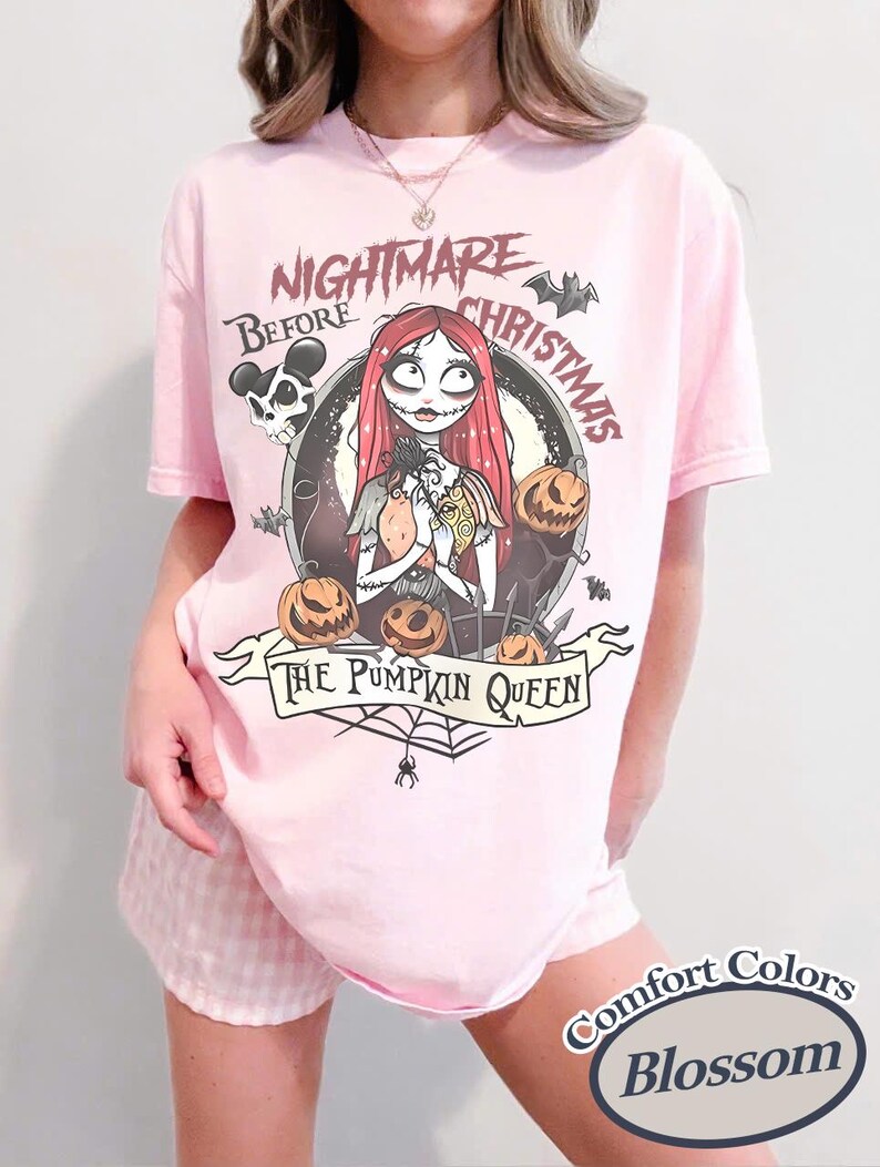 Puede incluir: Una camiseta rosa claro con un dise&ntilde;o de "Pesadilla antes de Navidad". El dise&ntilde;o incluye a Sally, calabazas, murci&eacute;lagos y el texto "Nightmare Before Christmas" y "The Pumpkin Queen". La camiseta est&aacute; etiquetada como "Comfort Colors Blossom".