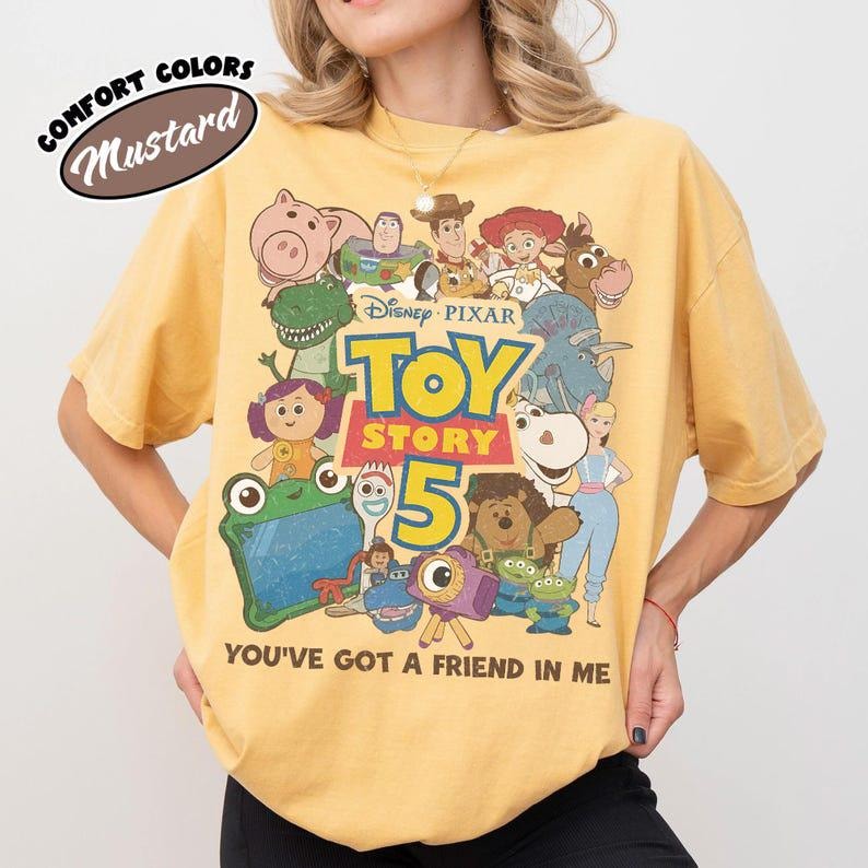 May include: A mustard yellow t-shirt featuring a graphic print of various Toy Story characters, the text "Disney Pixar TOY STORY 5", and the phrase "YOU'VE GOT A FRIEND IN ME". A small circular label in the top left corner reads "COMFORT COLORS MUSTARD".