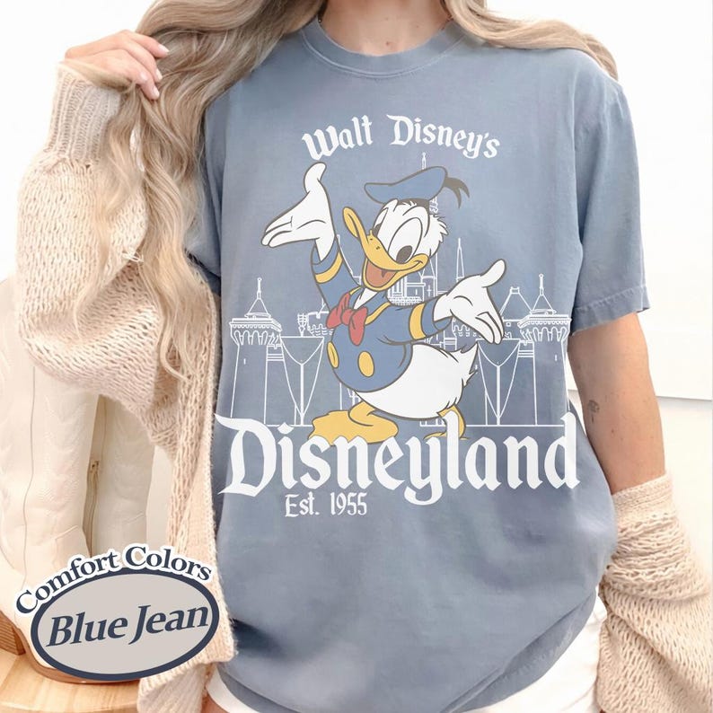 Puede incluir: Camiseta azul vaquero con un gr&aacute;fico de Pato Donald frente al castillo de Disneyland. El texto dice "Walt Disney's Disneyland Est. 1955". La camiseta es de la marca Comfort Colors.