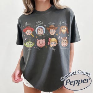 May include: Dark grey Comfort Colors t-shirt featuring cartoon character faces from the Toy Story movie. Characters include Jessie, Buzz Lightyear, Woody, Slinky, Alien, Hamm, Bo Peep, and Bullseye. The shirt has the text "Comfort Colors Pepper".