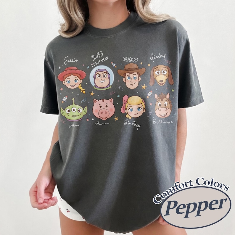 May include: Dark grey Comfort Colors t-shirt featuring cartoon character faces from the Toy Story movie. Characters include Jessie, Buzz Lightyear, Woody, Slinky, Alien, Hamm, Bo Peep, and Bullseye. The shirt has the text "Comfort Colors Pepper".