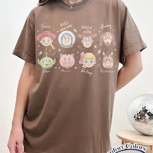 May include: Espresso-colored t-shirt featuring cartoon character portraits from a popular animated film. The shirt includes the names Jessie, Buzz Lightyear, Woody, Slinky, Alien, Hamm, Bo Peep, and Bullseye. The shirt also has the text "Comfort Colors" and "Espresso".