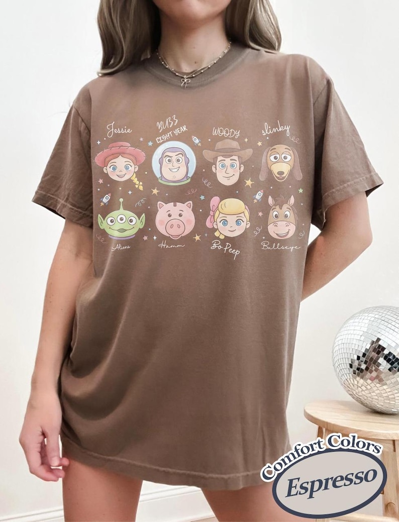 May include: Espresso-colored t-shirt featuring cartoon character portraits from a popular animated film. The shirt includes the names Jessie, Buzz Lightyear, Woody, Slinky, Alien, Hamm, Bo Peep, and Bullseye. The shirt also has the text "Comfort Colors" and "Espresso".
