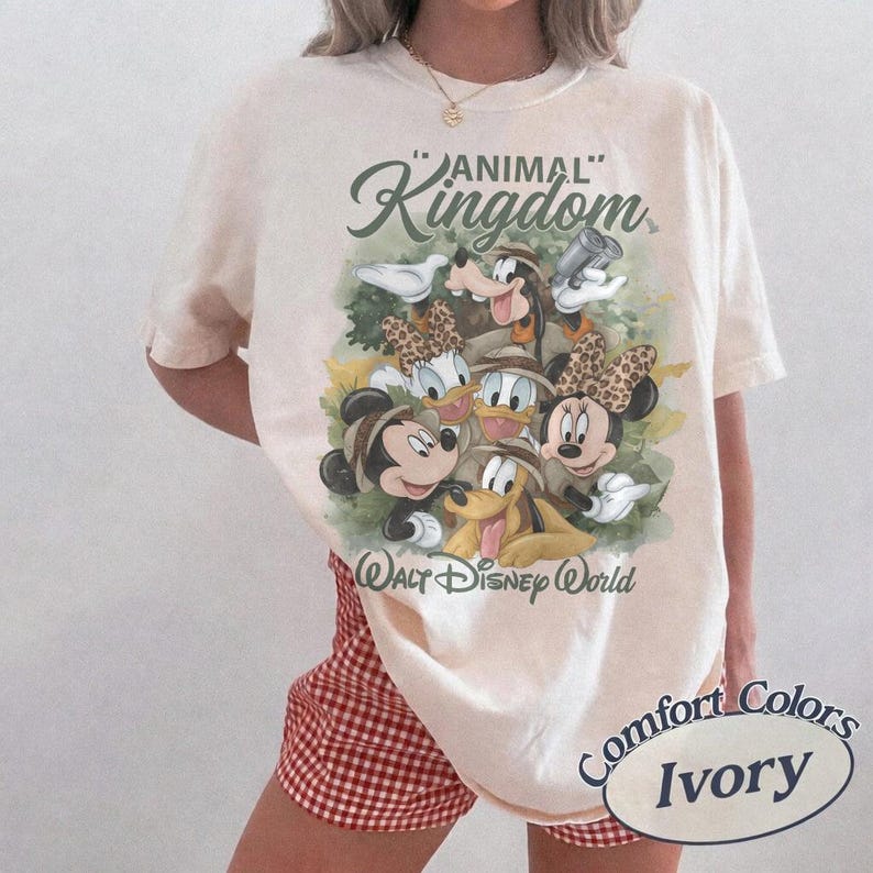 May include: Ivory-colored t-shirt featuring a graphic of Disney characters, including Mickey and Minnie Mouse, Donald Duck, and Goofy, with the text "Animal Kingdom" and "Walt Disney World". The shirt is a Comfort Colors brand.