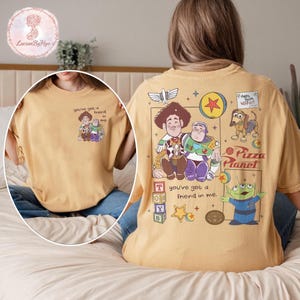 Puede incluir: Camiseta de color mostaza con un gr&aacute;fico de Woody y Buzz Lightyear con el texto "you've got a friend in me". La parte posterior de la camiseta muestra personajes y texto adicionales de Toy Story. Una camiseta divertida e informal.