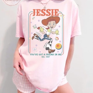 May include: Pink t-shirt featuring a cartoon graphic of Jessie from Toy Story. The design includes the name "JESSIE" in a retro font, the phrase "YOU'VE GOT A FRIEND IN ME," and the year "Est. 1997." The character is depicted in her signature cowgirl outfit.