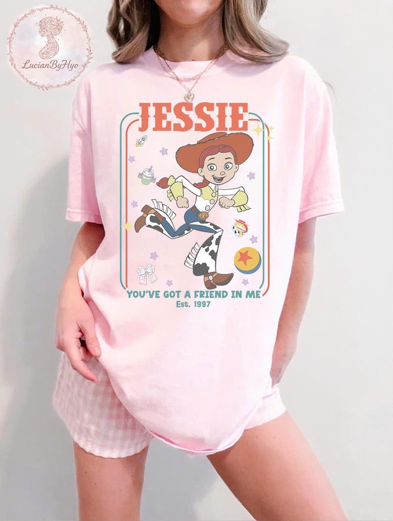 May include: Pink t-shirt featuring a cartoon graphic of Jessie from Toy Story. The design includes the name "JESSIE" in a retro font, the phrase "YOU'VE GOT A FRIEND IN ME," and the year "Est. 1997." The character is depicted in her signature cowgirl outfit.