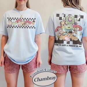 May include: Light blue t-shirt featuring Toy Story characters on the front and back. The front has a checkered border with characters, and the back displays a group of characters with the text "YOU'VE GOT A FRIEND IN ME". The shorts are red and white checkered.