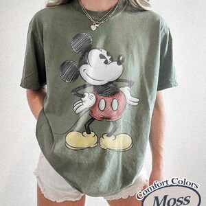 May include: Moss green Comfort Colors t-shirt featuring a sketch of Mickey Mouse in black, red, and yellow. The graphic shows Mickey with his hands on his hips. The shirt has short sleeves and a relaxed fit.