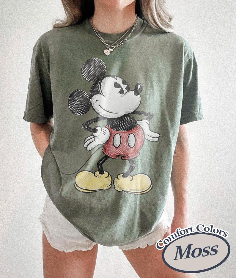 May include: Moss green Comfort Colors t-shirt featuring a sketch of Mickey Mouse in black, red, and yellow. The graphic shows Mickey with his hands on his hips. The shirt has short sleeves and a relaxed fit.