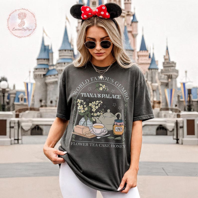 May include: Dark gray t-shirt with a graphic featuring the text "World Famous Gumbo, Tiana's Palace." The graphic includes a teapot, flowers, cake, honey, and teacup. The shirt is worn by a person wearing Minnie Mouse ears.