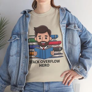 Könnte beinhalten: Beiges T-Shirt mit einer Cartoon-Illustration eines Mannes, der ein blaues Buch vor einem Stapel Bücher liest. Der Text "STACK OVERFLOW HERO" ist darunter gedruckt. Die Person trägt eine Jeansjacke.