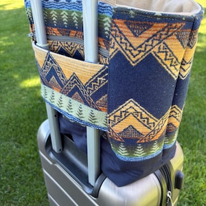 May include: A navy blue and tan tote bag with a geometric mountain pattern, attached to a silver rolling suitcase. The bag has a zippered pocket and tan handles. The background is green grass.