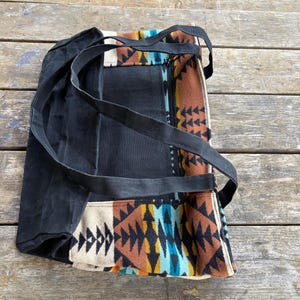 May include: Black canvas tote bag with long black straps. The bag features a colourful geometric pattern in shades of brown, orange, blue, and cream. The bag has a zip closure and is sitting on a wooden surface.