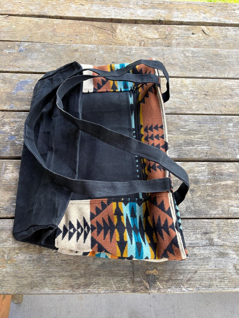 May include: Black canvas tote bag with long black straps. The bag features a colourful geometric pattern in shades of brown, orange, blue, and cream. The bag has a zip closure and is sitting on a wooden surface.