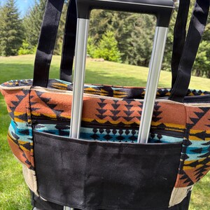 May include: A patterned tote bag with black straps and a black pocket, attached to a rolling suitcase. The bag features a colourful geometric design in shades of brown, orange, blue, and cream. The background shows a green lawn and trees.